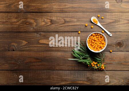 Bacche di fibbone di mare in ciotola, cucchiaio e foglie di pianta su tavola di legno. Composizione piatta, vista dall'alto. Foto Stock