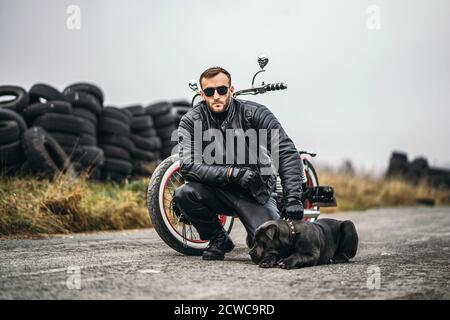 Biker in una tuta in pelle accovacciata vicino al suo cane e moto rosso sulla strada. Molti pneumatici sullo sfondo. Foto Stock