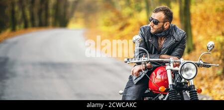 Uomo brutale con occhiali da sole e giacca in pelle seduto su una moto sulla strada nella foresta con sfondo colorato e blu. Foto Stock