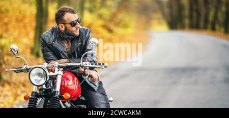 Uomo brutale con occhiali da sole e giacca in pelle seduto su una moto sulla strada nella foresta con sfondo colorato e blu. Foto Stock