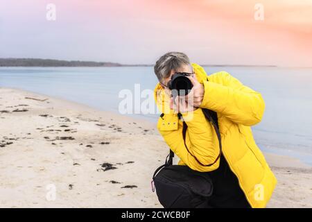 Fotografo maschile che scatta foto sulla spiaggia. Fotografo viaggiatore in giacca gialla guardando direttamente attraverso la fotocamera sulla costa del mare. Fotografia di viaggio Foto Stock