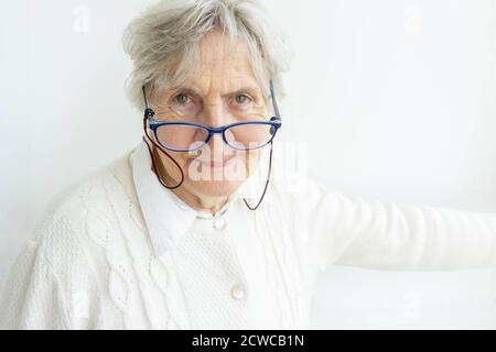 Ritratto di donna anziana che indossa occhiali sullo sfondo della parete. Vecchia donna in pullover bianco guardando la macchina fotografica. Grigio capelli donna anziana in occhiali isolare Foto Stock