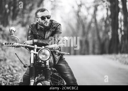 Uomo brutale con occhiali da sole e giacca in pelle seduto su una moto sulla strada nella foresta con sfondo colorato e blu. Foto Stock