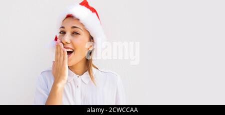 Giovane bella bionda caucasica con cappello Babbo Natale sorprendente con la mano davanti alla bocca con ampio sfondo bianco per la copia - Foto Stock