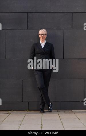 Ritratto di donna d'affari matura in abito nero appoggiato sul parete dell'edificio e sorridente a macchina fotografica all'aperto Foto Stock