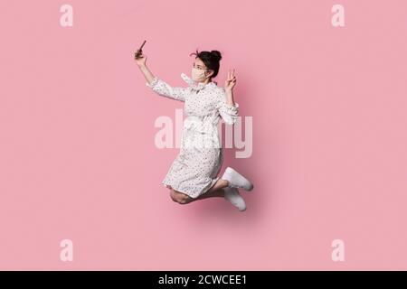 Donna bruna con maschera medica sul viso sta saltando dentro un abito estivo e fare un selfie su un rosa parete dello studio Foto Stock