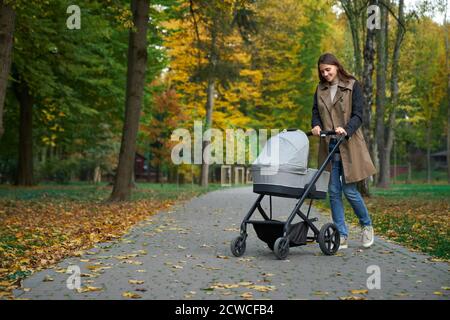 Donna elegante che cammina con passeggino grigio e bambino. Giovane madre che tiene il passeggino nelle mani e sorride al neonato. Concetto di camminare. Foto Stock