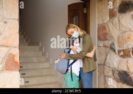 Donna caucasica e sua figlia entrano in casa, indossando maschere, in piedi accanto alla porta e abbracciando. Distanza sociale durante Covid 19 Coronaviru Foto Stock