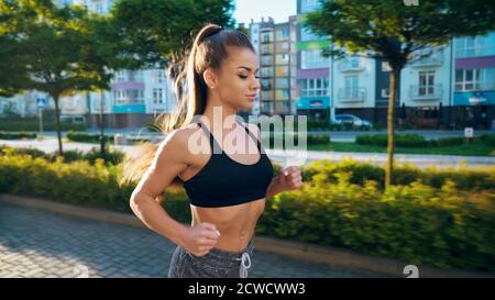 Vista laterale di una giovane bella ragazza runner indossando nero top jogging in estate mattina soleggiato in strade, edifici a più piani intorno.. Affascinante ragazza che pratica cardio allenamento all'aperto. Foto Stock