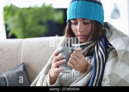 Giovane ragazza con influenza in cappello e sciarpa sotto le coperture tiene una tazza di tè nelle sue mani in appartamento Foto Stock