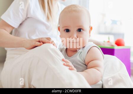 Neonato seduto alle ginocchia della madre. La famiglia si rilassa a casa. Capretto che guarda la macchina fotografica. Giorno della mamma Foto Stock