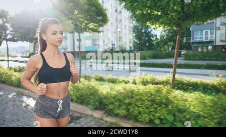 Vista laterale di una giovane runner femminile con outfit sportivo che fa jogging nella soleggiata mattinata estiva nelle strade. Affascinante fit ragazza che pratica cardio allenamento all'aperto, edifici a più piani e alberi intorno. Foto Stock