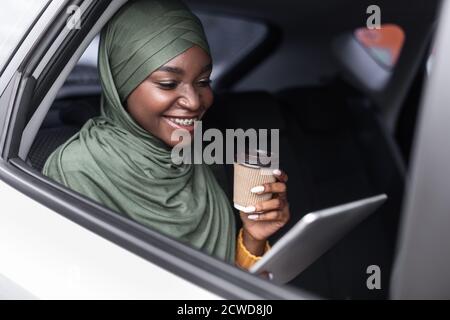 Donna musulmana nera che guida l'auto sul sedile posteriore con tablet digitale e caffè Foto Stock