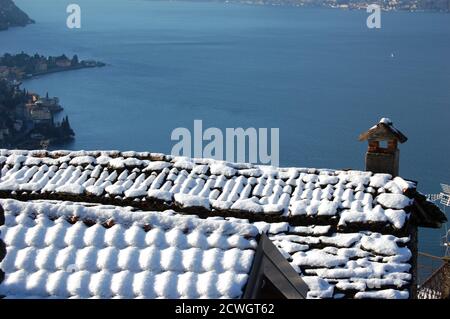 Vista panoramica invernale sulla costa del Lago di Como e su alcuni neve sul tetto di una casa e camino in primo piano Foto Stock