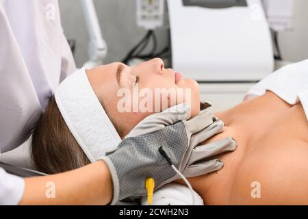 Hardware cosmetologo professionale touch viso cliente con guanti grigi microcorrente per la terapia della pelle miostimolante primo piano, vista laterale Foto Stock