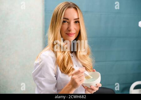 Bella donna sorridente medico di bellezza in bianco accappatoio tenere pennello e professionale peeling maschera bianca argilla in ciotola, procedura salone, trattamento della pelle Foto Stock