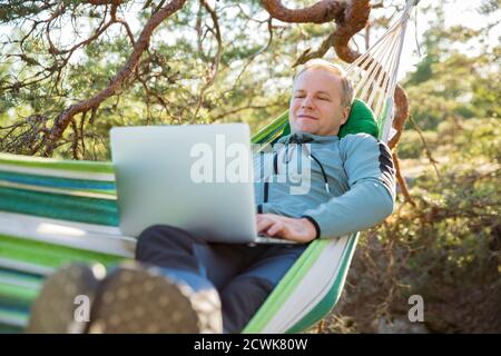 Un uomo che lavora su un computer portatile mentre si trova in un'amaca nei boschi. Autoisolamento, freelancing, lavoro remoto e distancing. Paesaggio scandinavo Foto Stock
