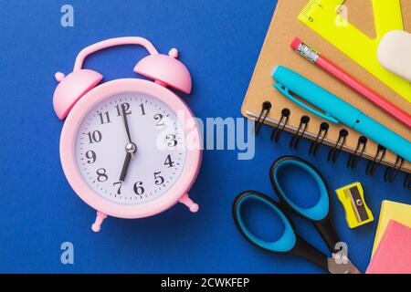 Sveglia e forniture scolastiche su sfondo blu, vista dall'alto. Concetto sul tema della preparazione alla scuola. Foto Stock