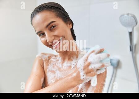 Giovane donna di charme che usa loofah esfoliante mentre si prende la doccia Foto Stock