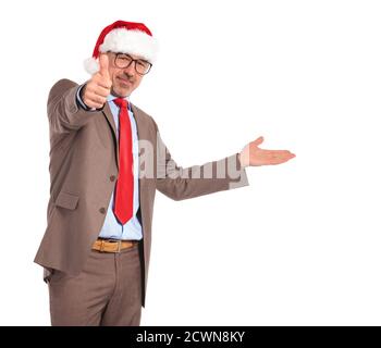 uomo d'affari vecchio che indossa il cappello di babbo natale che presenta e fa il ok pollici su segno mano su sfondo bianco Foto Stock