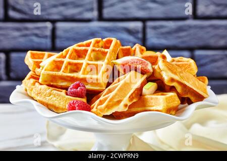 Waffle belgi con frutti di bosco freschi, pere, fichi su una torta stand su un tavolo di marmo bianco con un muro di mattoni sullo sfondo Foto Stock