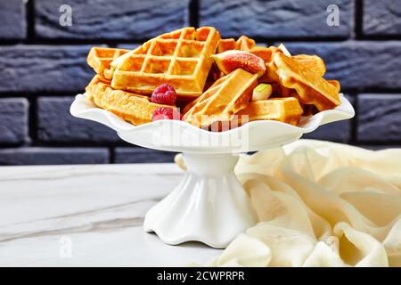 Waffle belgi con frutti di bosco freschi, pere, fichi su una torta stand su un tavolo di marmo bianco con un muro di mattoni sullo sfondo Foto Stock