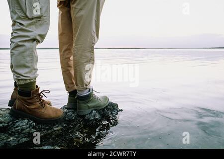 Primo piano di una coppia irriconoscibile in stivali da trekking in piedi sulla roccia e contemplando il paesaggio acquatico Foto Stock