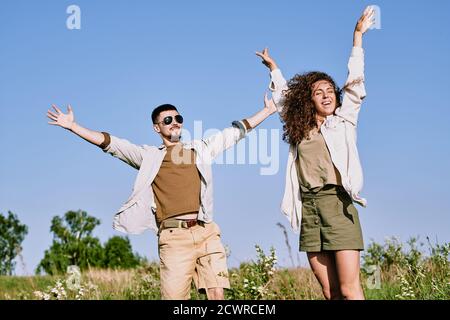 Giovane coppia allegra ed eccitata in comoda vestita in piedi con rialzato armi mentre ti godi un viaggio in campagna Foto Stock