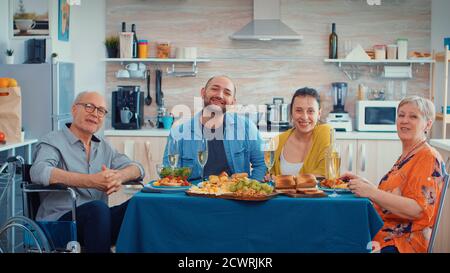 Famiglia Extanted e nonno immobilizzato che ha una videochiamata nella loro moderna, cucina di lusso durante la cena. POV online internet moderno, chat, comunicazione, chat conversazione chiamata via webcam. Foto Stock