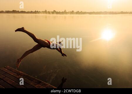 Giovane uomo attivo che salta in acqua dal molo di legno Foto Stock