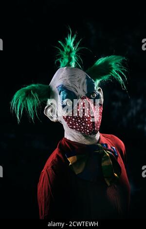 closeup di un clown malvagio inquietante, con i capelli verdi, indossando un costume rosso sporco e una cravatta d'arco sporca, indossa una maschera facciale rossa modellata con puntini bianchi, Foto Stock