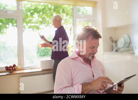 Uomo anziano che utilizza un tablet digitale in una cucina soleggiata al mattino Foto Stock