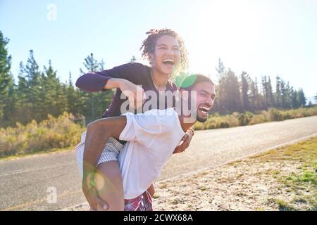 Felice giovane coppia giocosa piggybacking sulla soleggiata strada estiva Foto Stock