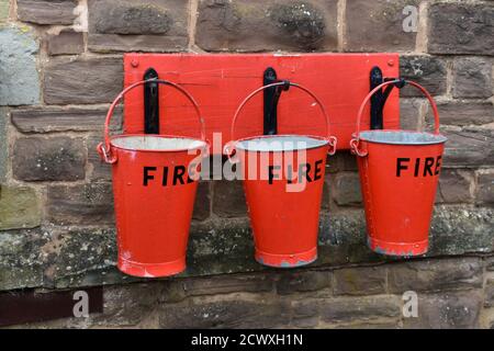 Primo piano di 3 secchi antincendio rossi appesi ai ganci su un muro di pietra all'esterno Foto Stock