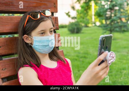 ragazza in maschera protettiva che tiene uno smartphone. ragazza teen che si siede su panchina in verde parco con telefono cellulare. Prima fase di allentamento del coronavirus Foto Stock