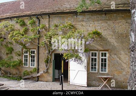 Una porta si apre sulla strada verso un granaio di pietra Cotswold. La glicine cresce oltre la porta Foto Stock