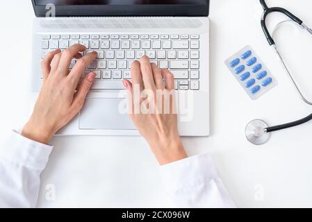 vista dall'alto delle mani di un medico caucasico che usa un computer portatile moderno sulla parte superiore di una scrivania bianca Foto Stock