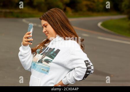 Shenandoah Valley, VA, USA 09/27/2020: Una giovane donna ispanica con capelli marroni e felpa con cappuccio sta scattando una foto con uno smartphone. Ha un sorriso su h. Foto Stock