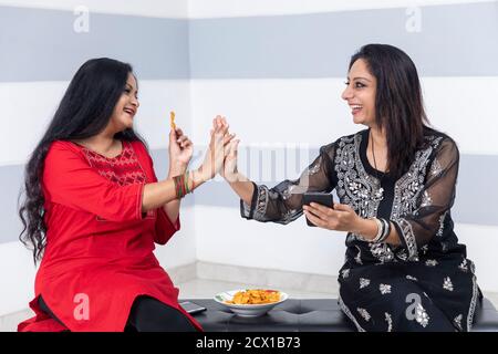 Ritratto di due casalinghe indiane che mangiano patatine e si divertono, dando five alte, sorridendo e godendosi insieme a casa. Foto Stock