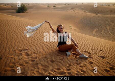 Felice donna bionda sorridente siediti nella sabbia del deserto con una camicia bianca frullato sul vento, goditi un tour safari a Dubai, Emirati Arabi Uniti, turista in Medio Oriente, fuoristrada A. Foto Stock