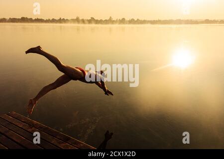 Giovane uomo attivo che salta in acqua dal molo di legno Foto Stock