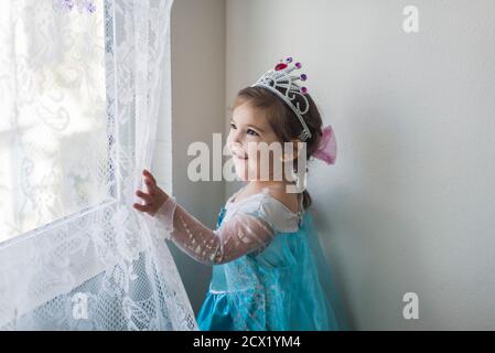 Sorridente ragazza in costume principessa e tiara che tocca pizzo bianco tenda Foto Stock