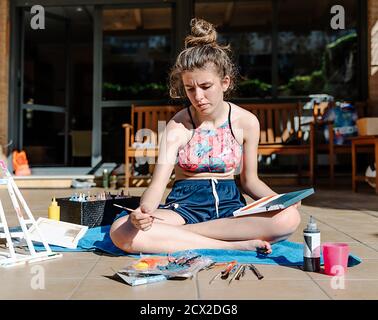 ragazza bianca che dipinge un'immagine seduta su una terrazza con tutti gli elementi di pittura intorno come pennelli, vernici, acqua,... Foto Stock