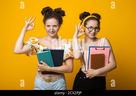 I gemelli con capelli ricci stanno gesturando il segno giusto che indossa gli occhiali con cuffie e cartelle da scuola su un giallo parete dello studio Foto Stock