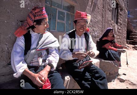 Due uomini che ridono a chiacchierare e a lavorare a maglia cappelli. la plaza, l'isola di Taquile, il lago Titicaca, Perù. Una donna locale si siede dietro, filando lana con un fuso di goccia. Foto Stock