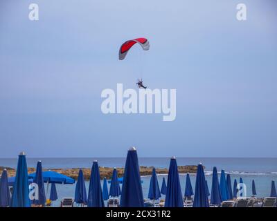 Parapendio motorizzato con ala rossa che vola in alto nel cielo blu serale sopra la spiaggia di Nissi con ombrelloni blu ad Ayia Napa, Cipro. Foto Stock