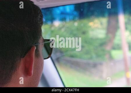 Un uomo caucasico con i capelli neri che indossano gli occhiali da sole messo a fuoco sopra La strada durante la guida Foto Stock
