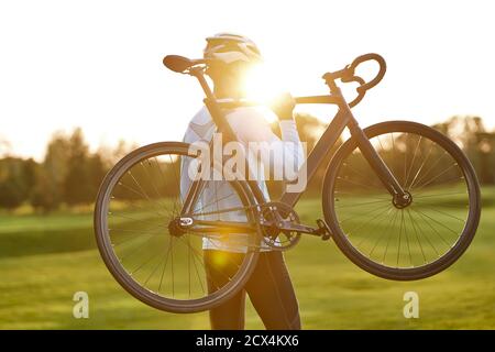Forte atletico uomo in abbigliamento sportivo che tiene in mano una bicicletta mentre si trova nel parco al tramonto, in bicicletta all'aperto e godendo di una splendida vista sulla natura. Stile di vita attivo e sport Foto Stock