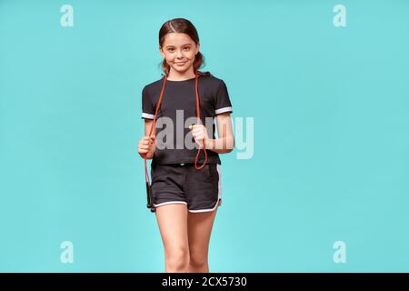 Bambini impegnati nello sport. Ragazza adolescente carina in nero sport con corda salto guardando la fotocamera e sorridendo mentre si posa isolato su blu Foto Stock