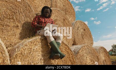 Agricoltore nero africano seduto sul pagliaio e lavorando sul portatile. Scatto ad angolo basso. Foto di alta qualità Foto Stock